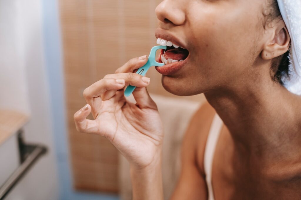 A young black girl stands in front of a bathroom mirror with a towel wrapped around her head. She is holding a piece of dental floss between her fingers and is in the process of flossing her teeth taught by Aspen Dental Care, a Calgary dentist. The bathroom is brightly lit, and a shower can be seen in the background.