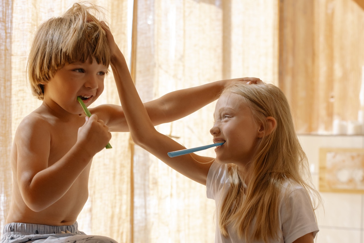 Two children with big smiles stands on a stool in front of a bathroom sink. They are holding a toothbrush with a dollop of toothpaste on it, and their other hand is holding onto the counter for balance. In the background, a colorful toothbrush holder can be seen, with toothbrushes of various sizes and colors.